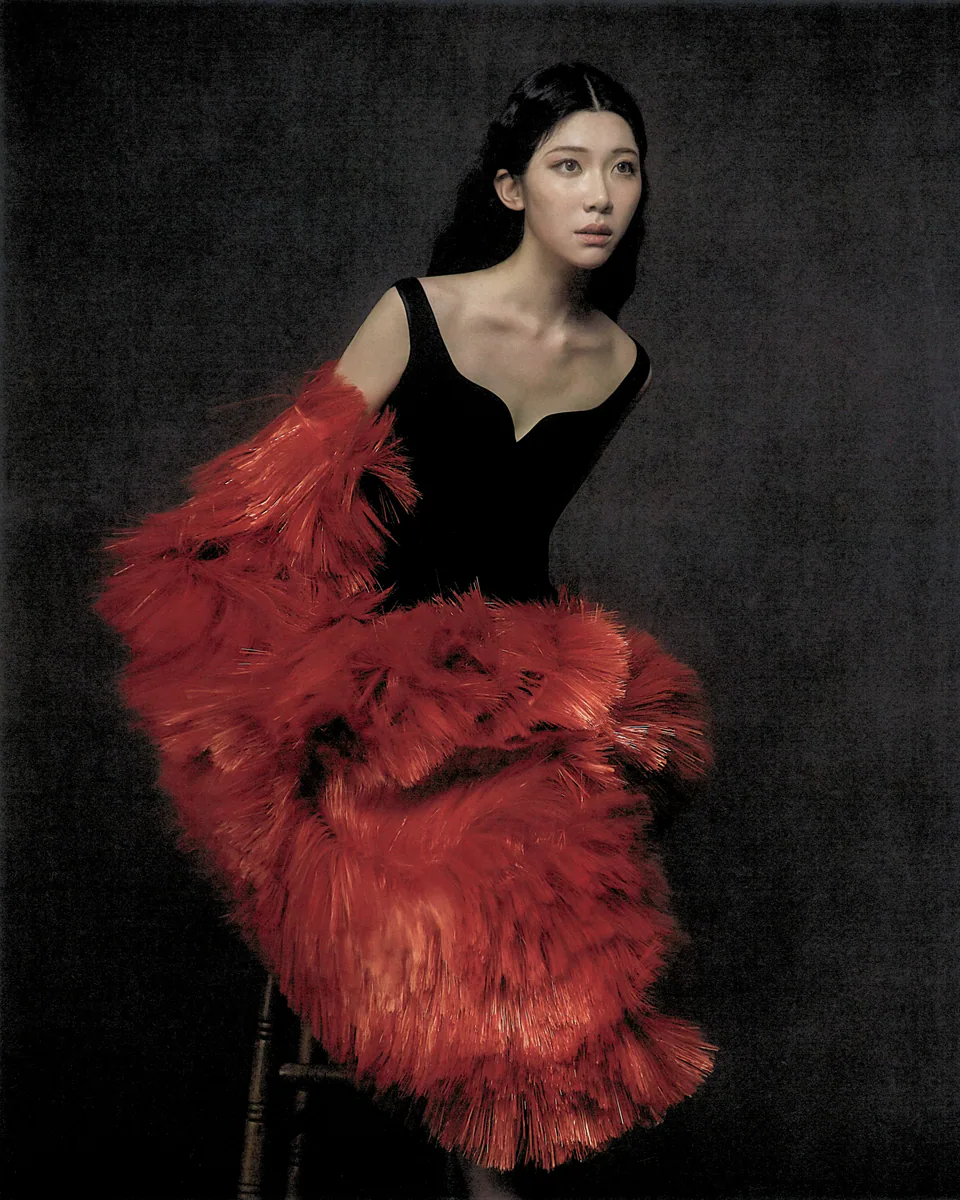 Sammi Chen seated in black with a red feather wrap against charcoal.